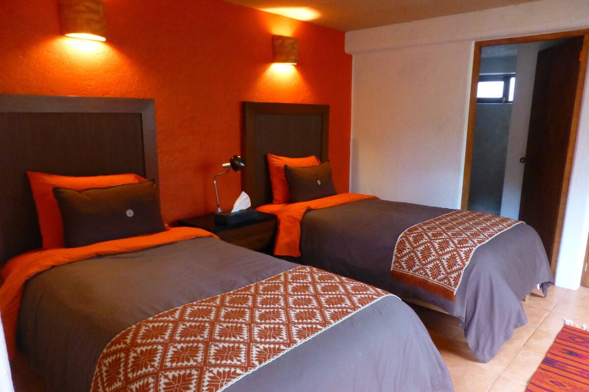 A hotel room with two beds, each with a patterned bedspread and pillows. There are wall-mounted lights above the beds and a door leading to another room.