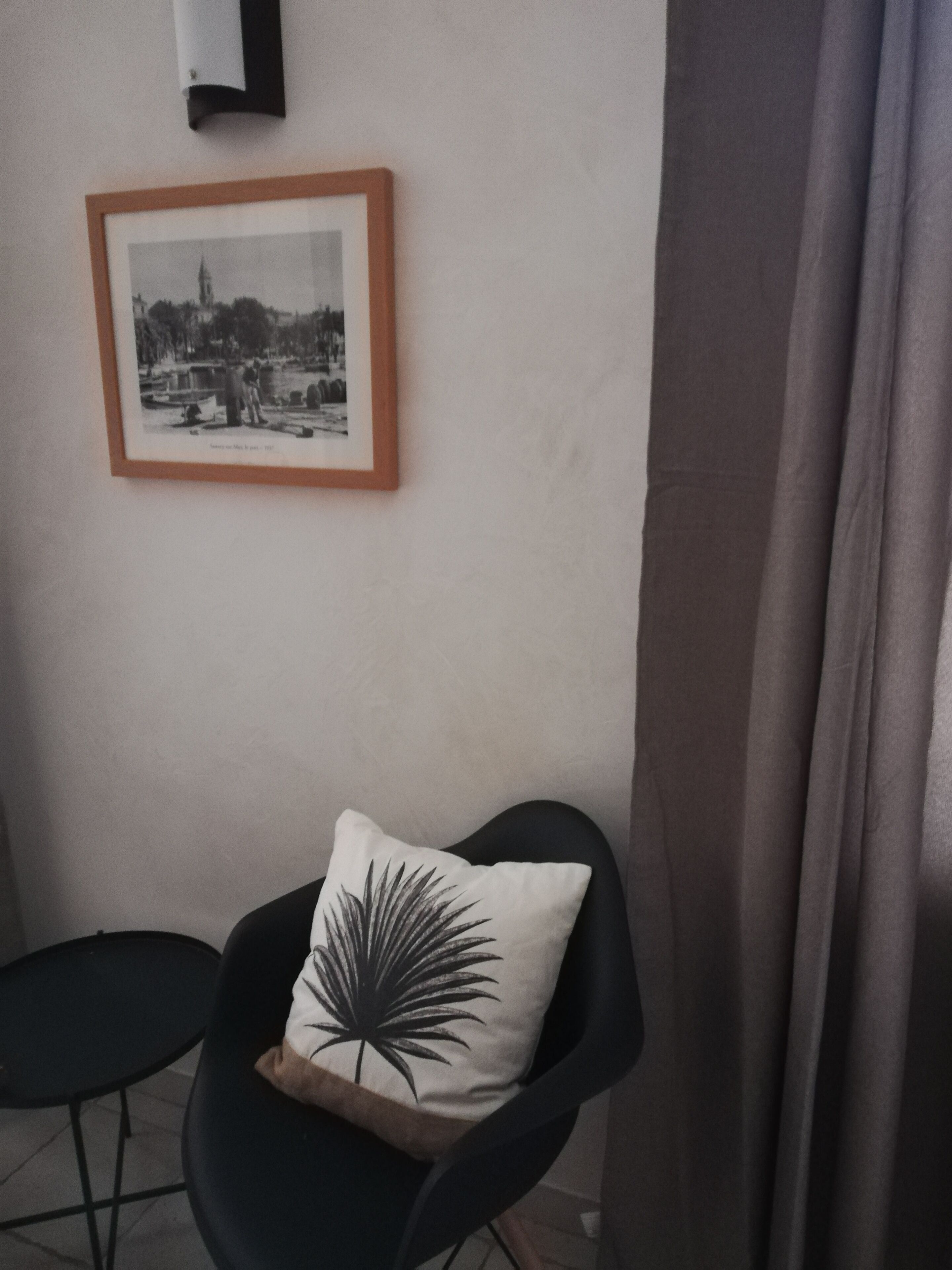 Una habitación pequeña con una silla, una mesa, un cuadro en la pared y una cortina.