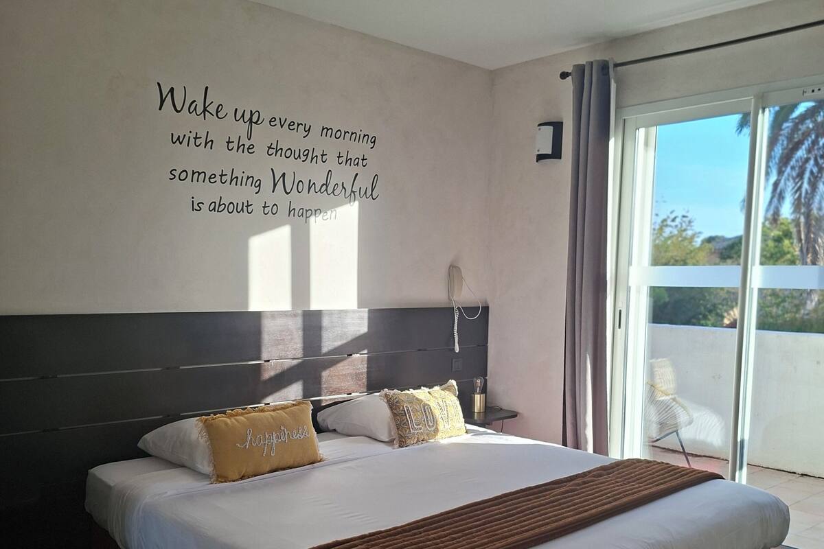 Ett hotellrum med en stor säng, ett inspirerande citat på väggen och utsikt över palmerna genom fönstret.