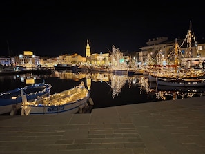 Un puerto deportivo de noche con barcos amarrados e iluminados por luces colgantes, una torre destacada al fondo y una superficie de agua tranquila que refleja las luces.