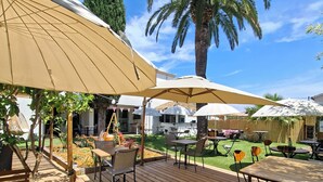 An outdoor patio with wooden tables and chairs, umbrellas, and a palm tree. - Hôtel Synaya (Sanary-sur-Mer)