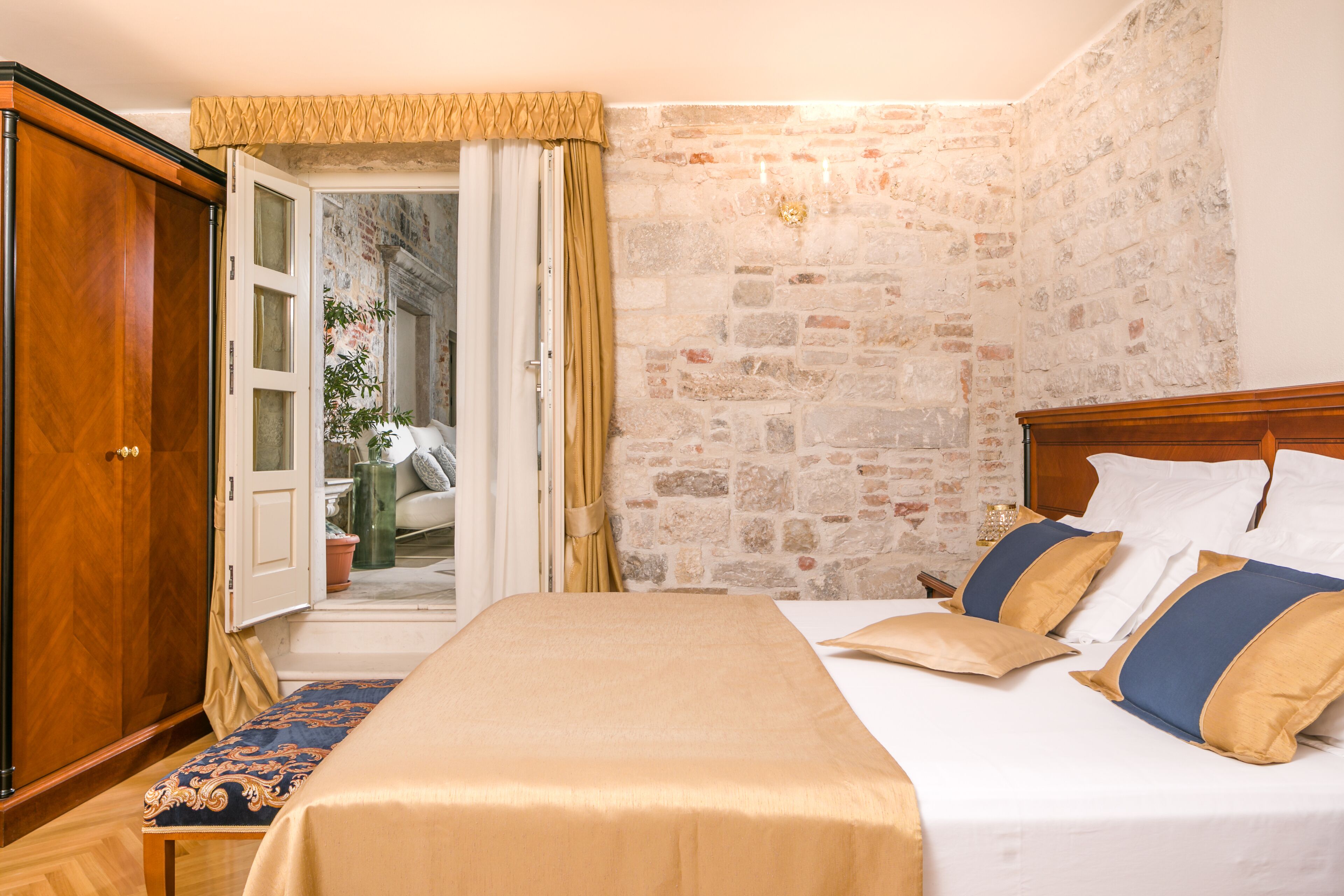 A bedroom with a stone wall, a wooden wardrobe, a bed with beige and blue bedding, and a bench.