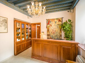 Un comptoir d’accueil en bois surmonté d’un plateau en marbre, un lustre, une grande tapisserie et une plante en pot.
