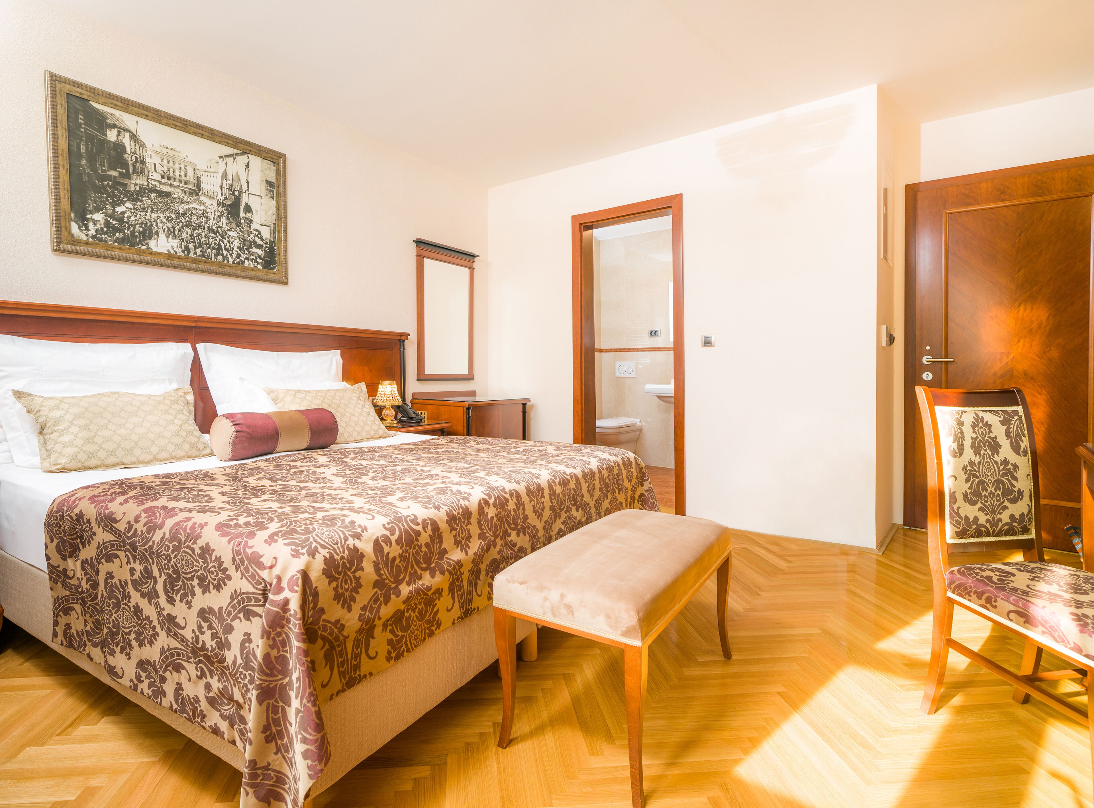 A hotel room with a large bed, a wooden bench, a chair, a bedside table with a lamp, a mirror, and a framed picture on the wall.