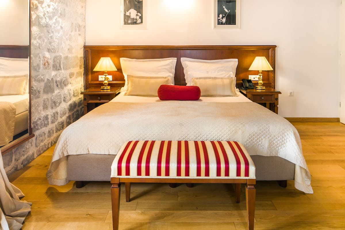 A neatly made bed with white linens and a red pillow, flanked by two bedside tables with lamps, in a room with wooden flooring and a stone accent wall.
