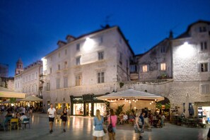 Une place historique dotée d’une terrasse, bordée d’immeubles en pierre, et où se trouve un café appelé « Palaça Judita ».