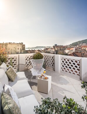 Une terrasse sur le toit, avec un fauteuil en osier blanc, un canapé blanc et une petite table sur laquelle sont posés une boisson et des pâtisseries. La terrasse offre une vue sur le paysage urbain, avec des bâtiments et un pont qui se dessinent à l’horizon.
