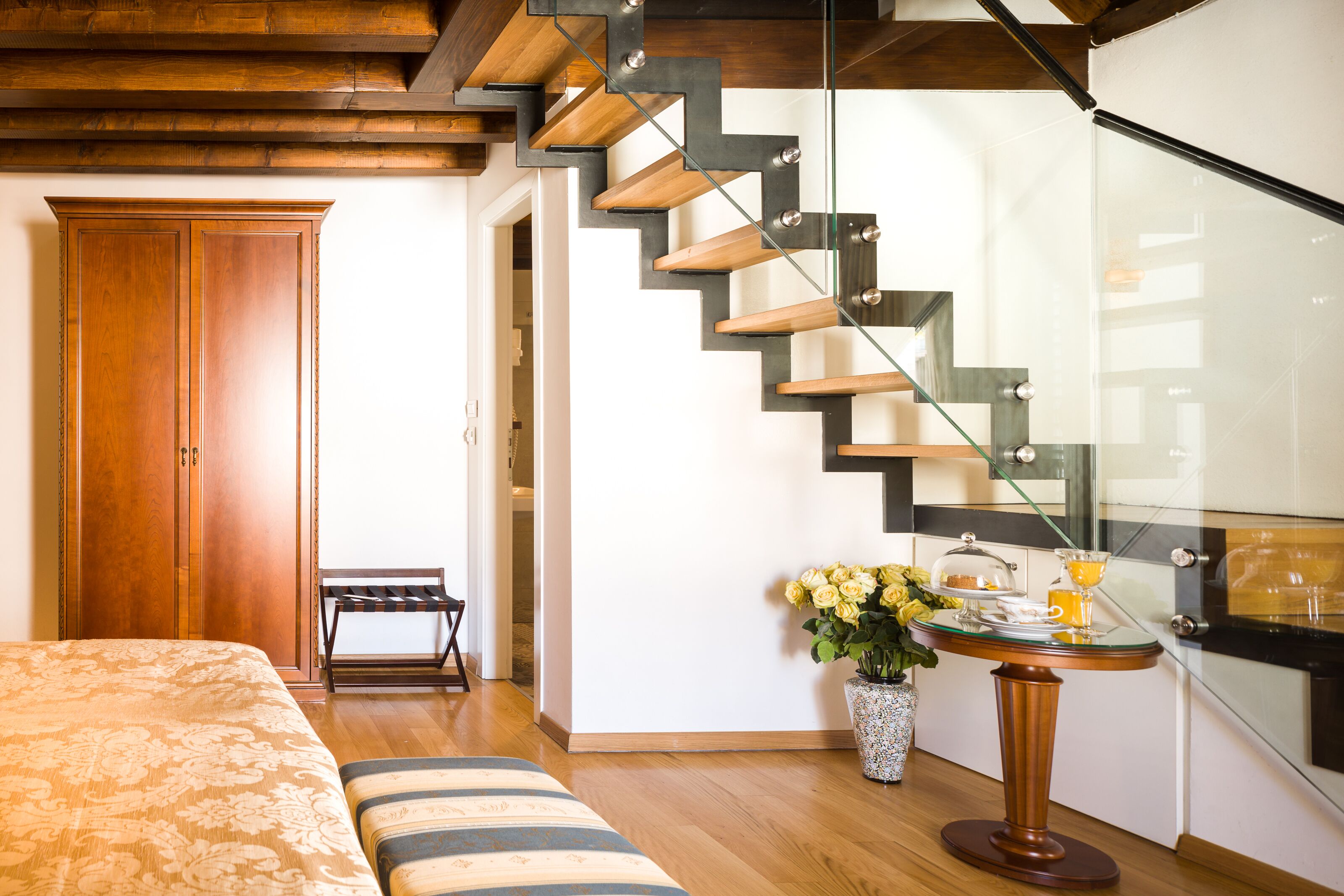 Ein modernes Schlafzimmer mit einer Holztreppe, einem Bett mit floralem Überwurf, einer Bank und einem kleinen Tisch mit Blumen und einem Glas.