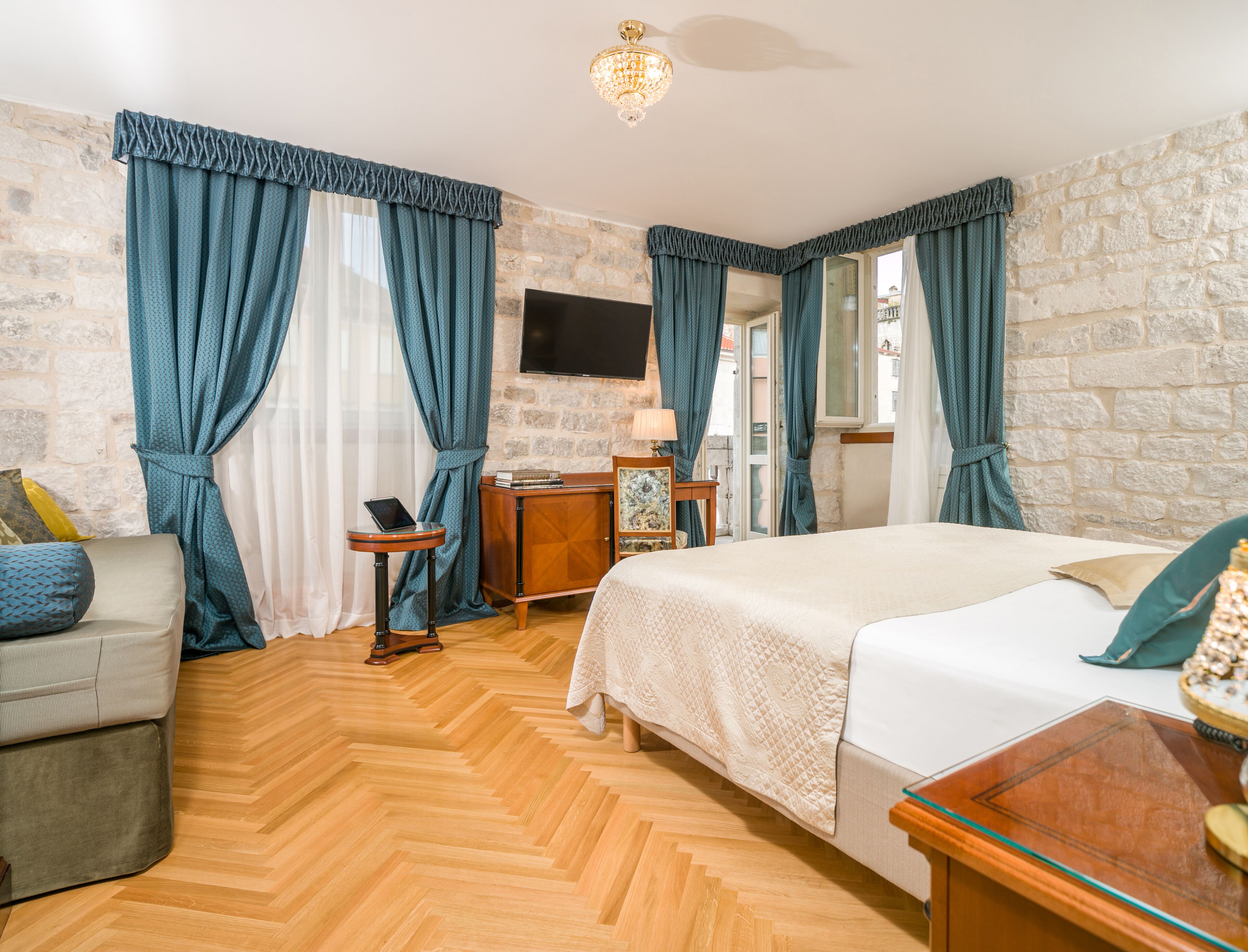 Quarto de hotel com piso de madeira em padrão herringbone, cama com colcha branca, sofá com almofada estampada, mesa de cabeceira de madeira e TV fixada na parede.
