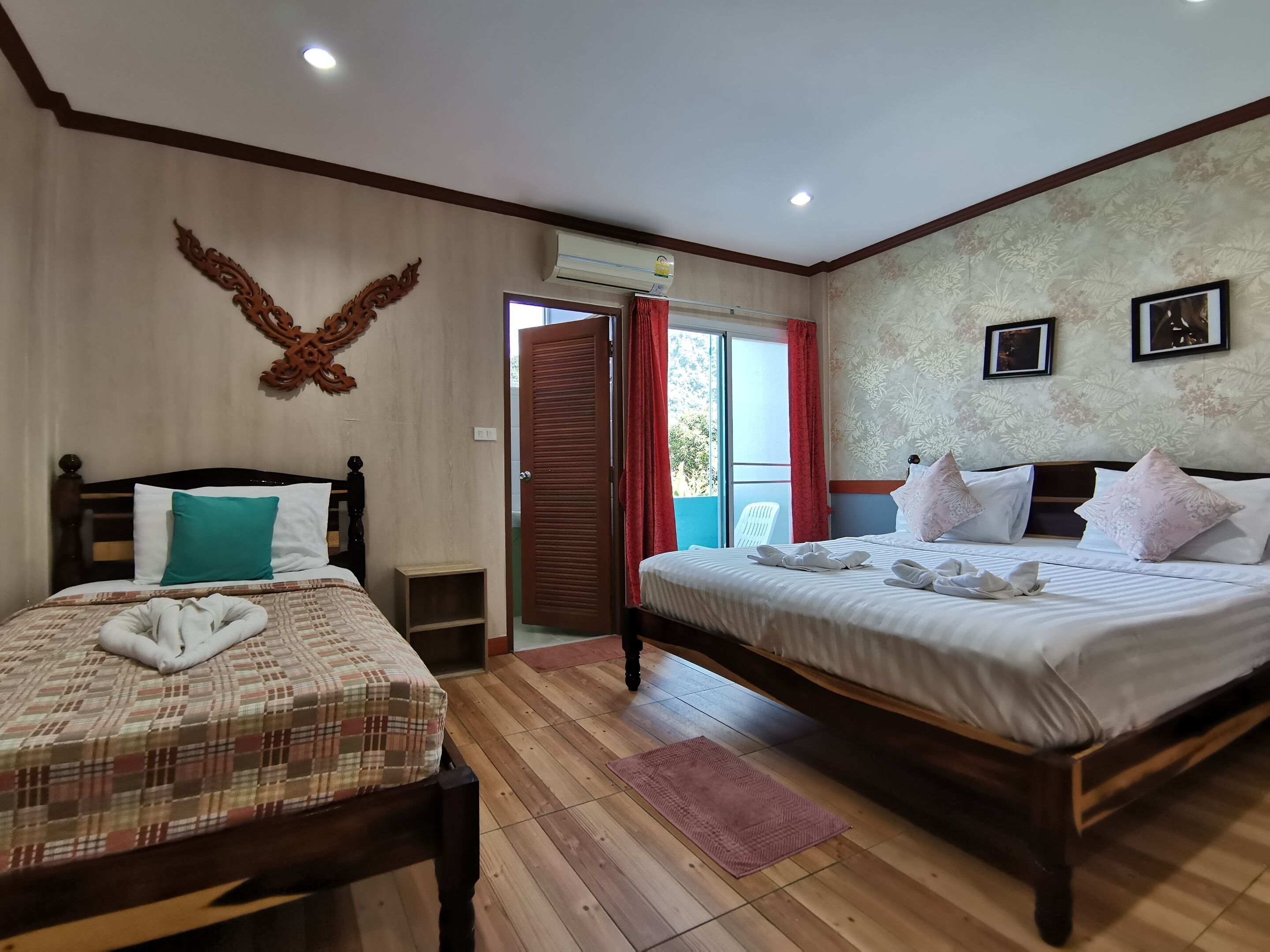 A hotel room with two beds, a wooden floor, a large window with red curtains, and a wall-mounted air conditioner.
