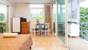 A hotel room with a bed, a desk, a refrigerator, a dining table with chairs, and a balcony with a view of greenery. - Kamala Beach Residence (Kamala)