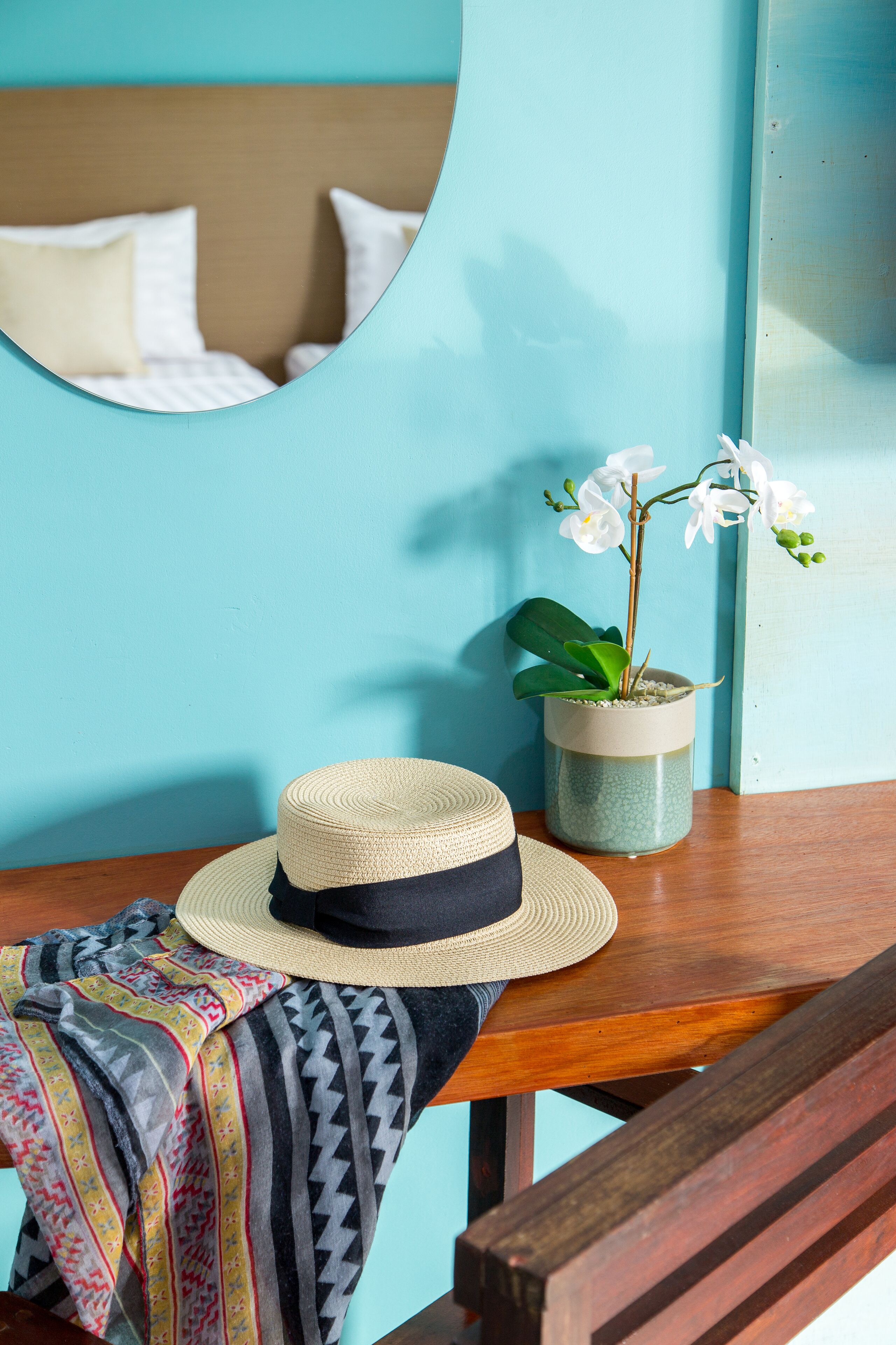 A room with a wooden headboard, a mirror, a potted orchid, and a straw hat on a wooden table.