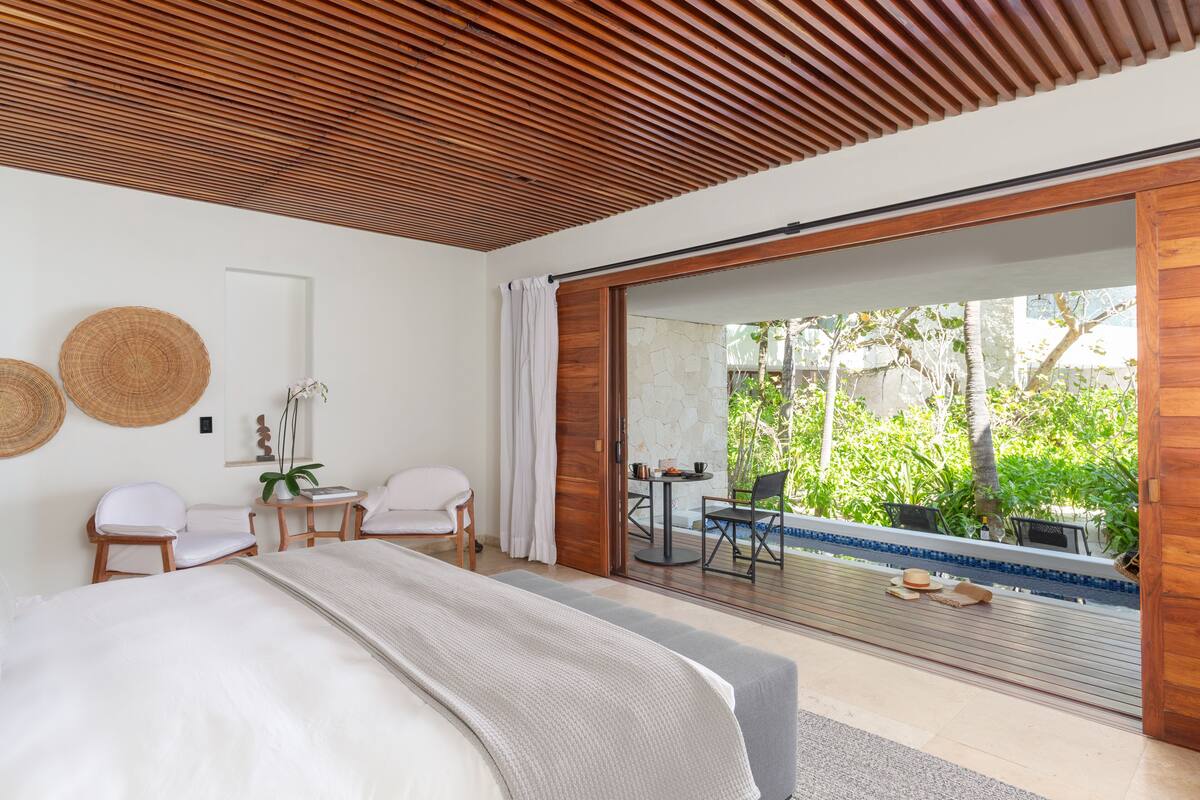 Un dormitorio moderno con una cama grande, dos sillas blancas, una mesita y vistas a una piscina y zonas verdes al exterior.