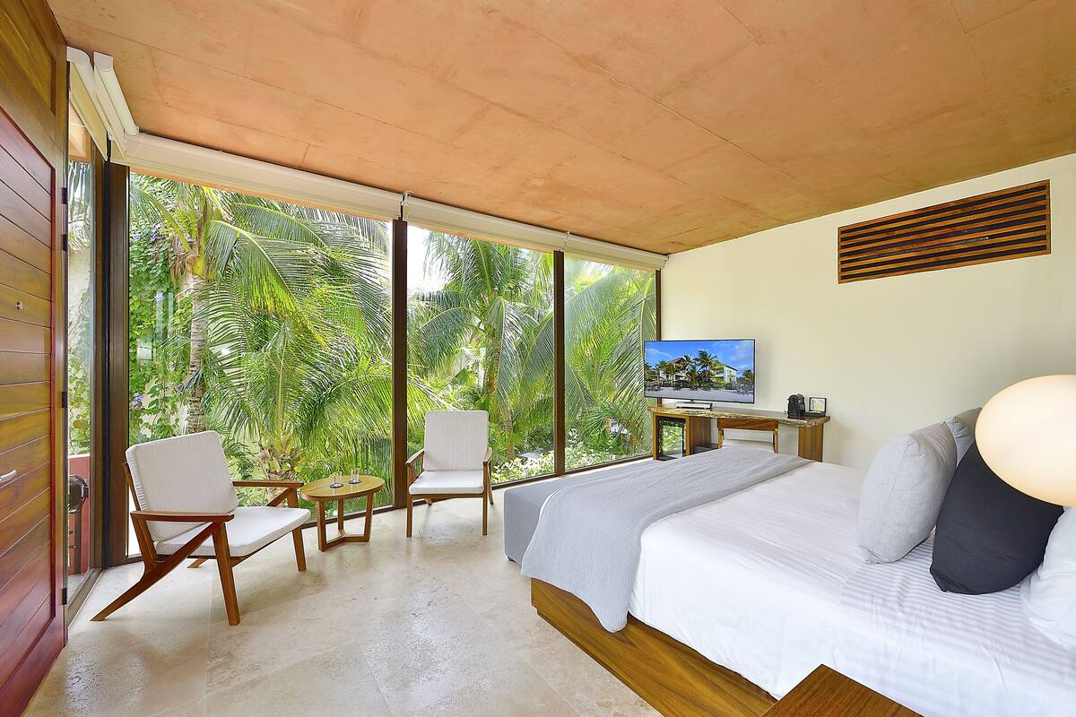 Un dormitorio con una cama grande, televisión, escritorio y vistas a abundante vegetación a través de amplios ventanales.