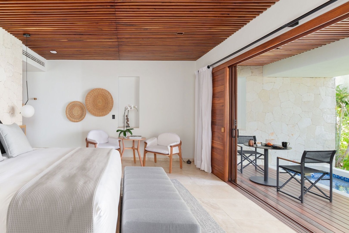 Une chambre à coucher comprenant un lit, des tables de chevet, un bureau, des chaises et donnant sur un espace extérieur avec une piscine.