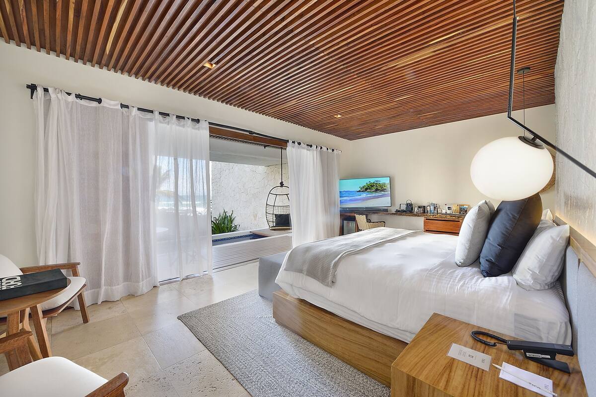 Una habitación de hotel moderna con techo de madera, una cama grande con almohadas, un escritorio con silla, un televisor y vistas al exterior.