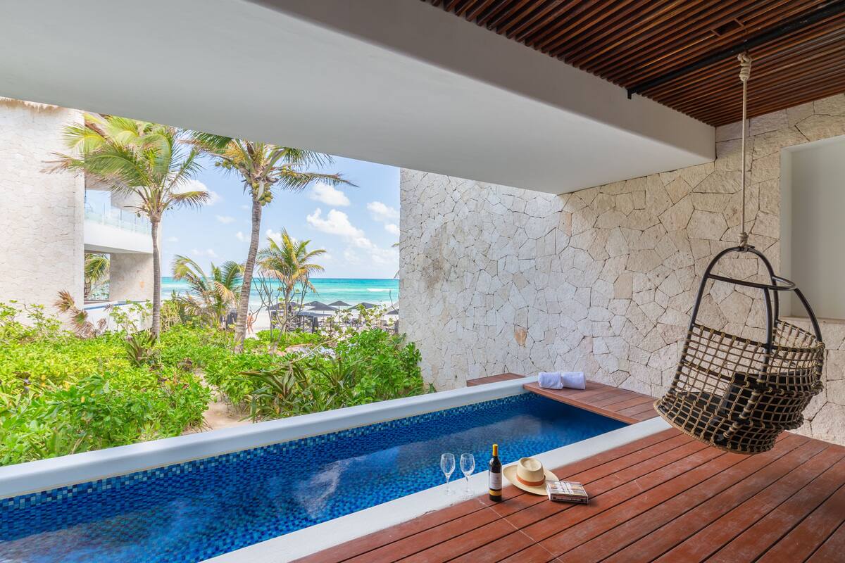 Terraza de madera con piscina, una silla colgante y vistas al océano y palmeras.