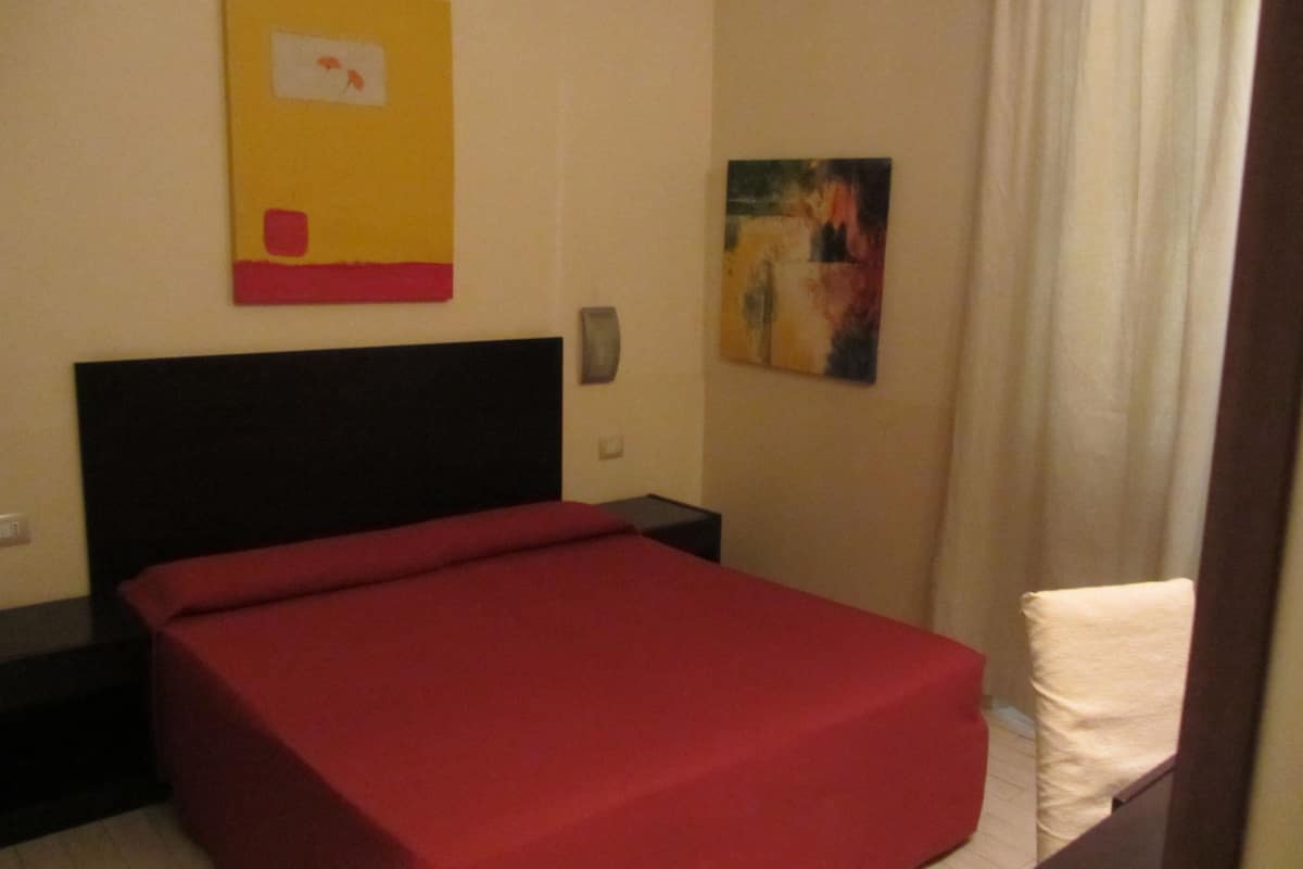 A room with a bed, a nightstand, a wall-mounted light, and a painting.