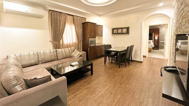 Ein modernes Wohnzimmer mit Sofa, Couchtisch, Esstisch und Stühlen. Es gibt eine Küchenzeile mit Schränken und einer Mikrowelle. Der Raum hat einen Holzboden und eine Deckenleuchte.