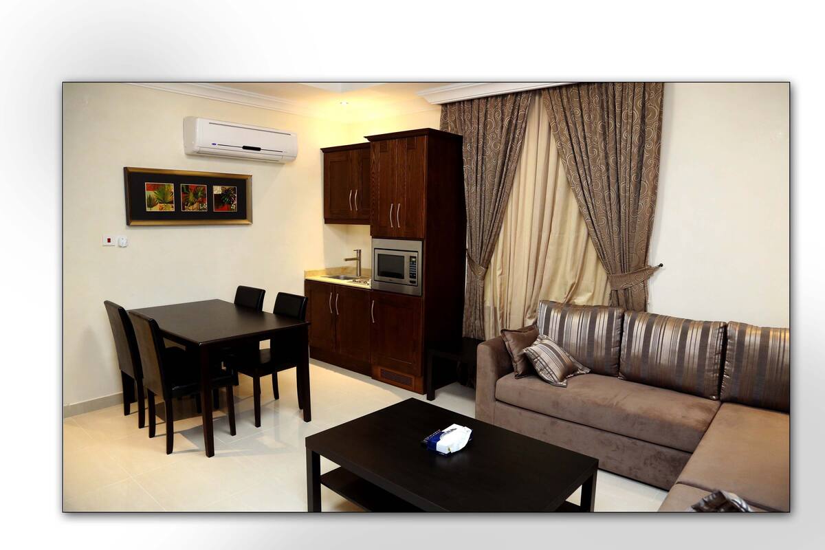 A living room with a sofa, a dining table with chairs, a kitchen area with cabinets, and an air conditioning unit.