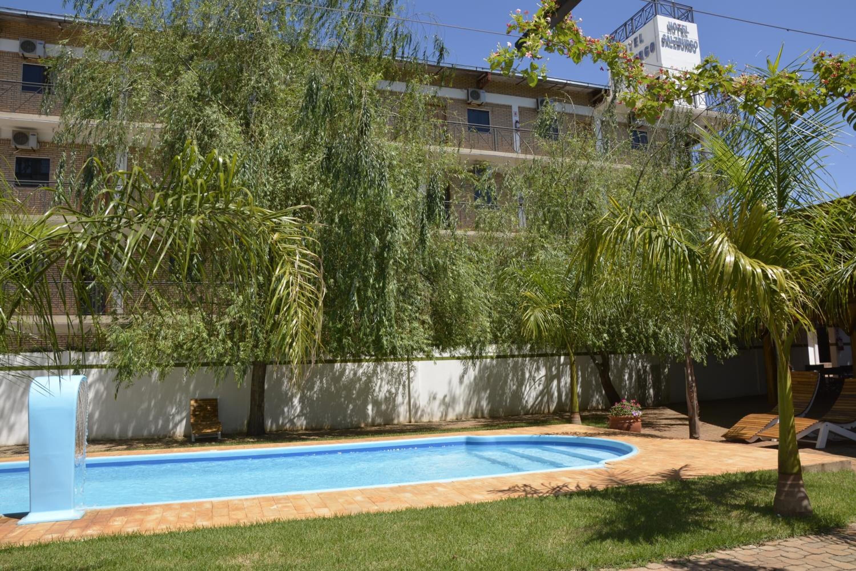 Une piscine entourée d’une terrasse, un mur blanc et un bâtiment avec des balcons en arrière-plan.