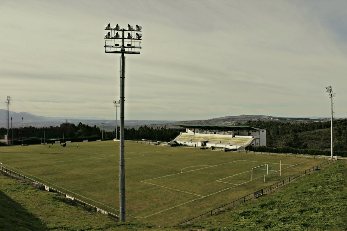 Um campo desportivo amplo e aberto, com balizas, um alto mastro de iluminação e um edifício com arquibancadas ao fundo.