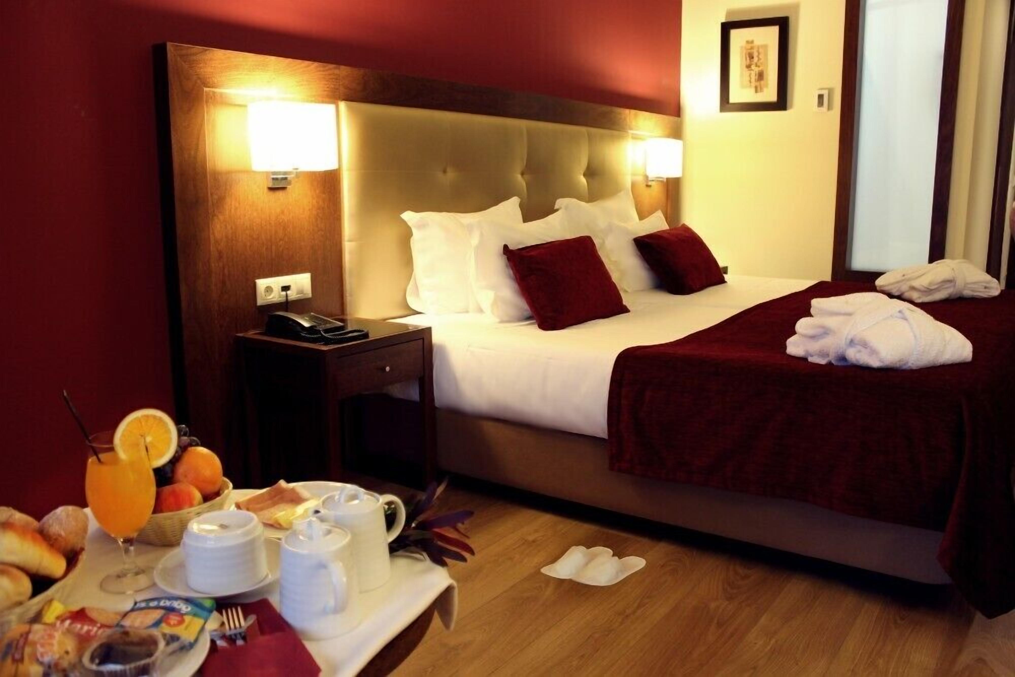 A hotel room with a large bed, two bedside lamps, a nightstand with a phone, and a breakfast tray with orange juice, croissants, and coffee.