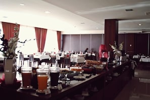 Uma área de refeições de hotel com várias mesas preparadas para o pequeno-almoço, incluindo diversos pratos e bebidas.