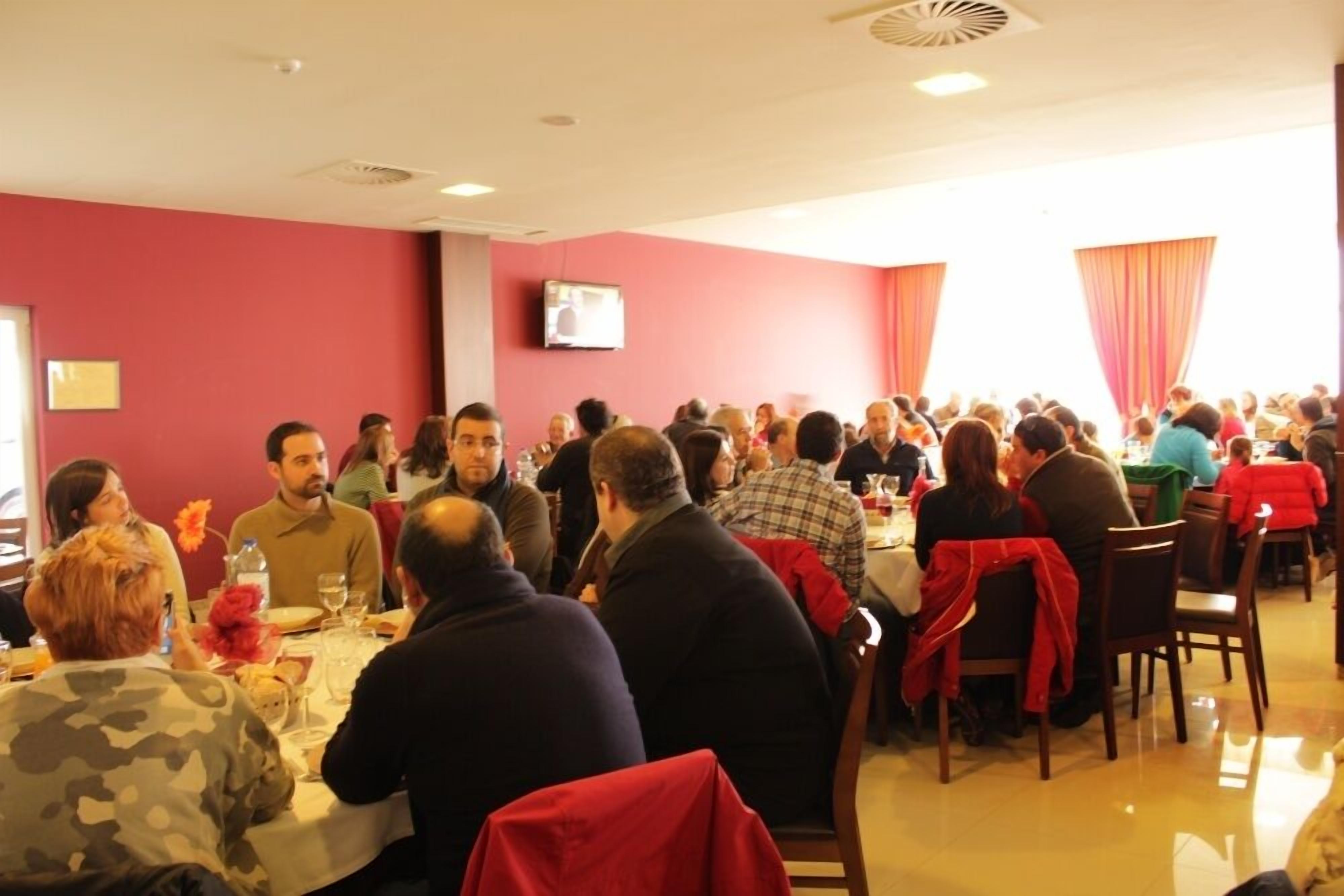 A crowded dining area with tables and chairs, a television mounted on the wall, and a window with curtains.