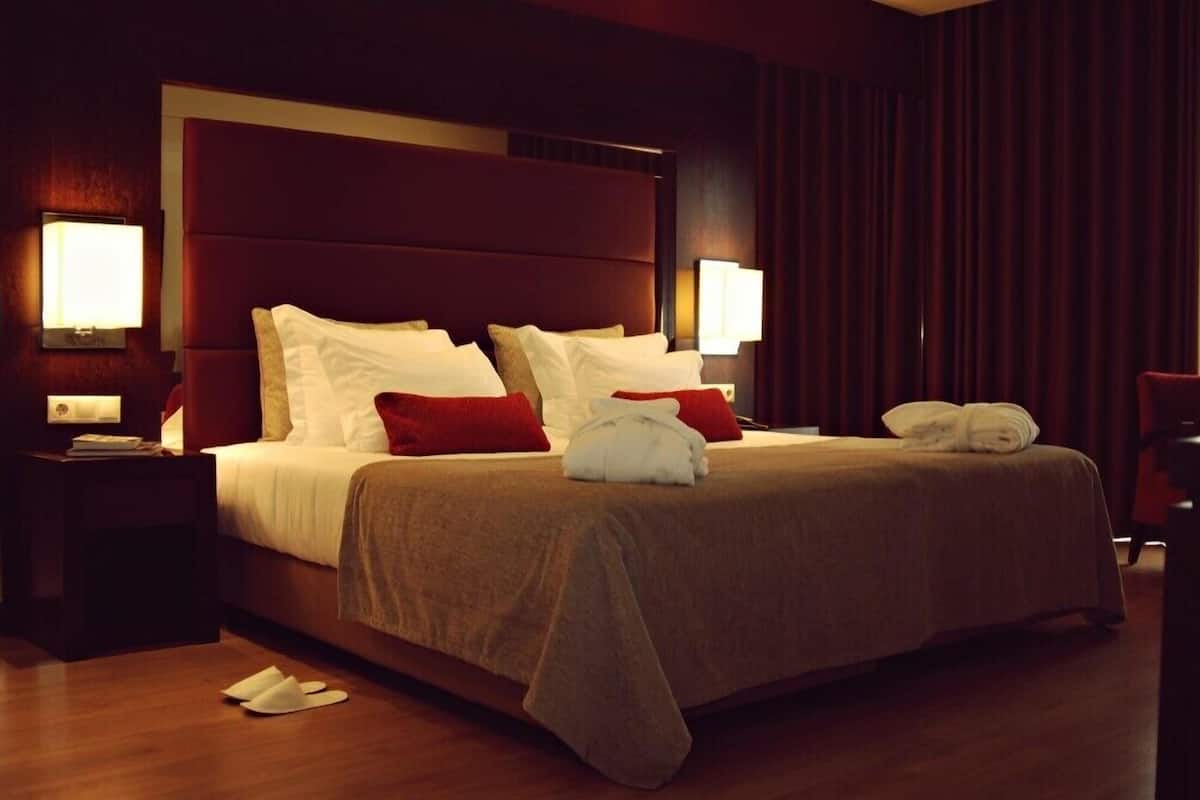 Una cama bien tendida con almohadas blancas y rojas, una colcha marrón y una toalla doblada al lado. Hay dos mesitas de noche con lámparas.