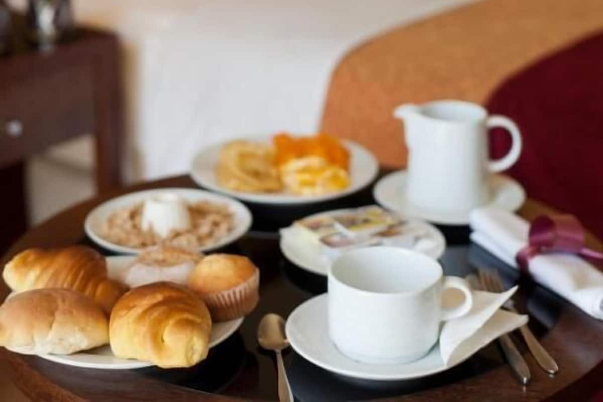 Una mesa de desayuno ordenada con cruasanes, pasteles y una cafetera, con vistas al dormitorio de un hotel que incluye una cama y almohadas.