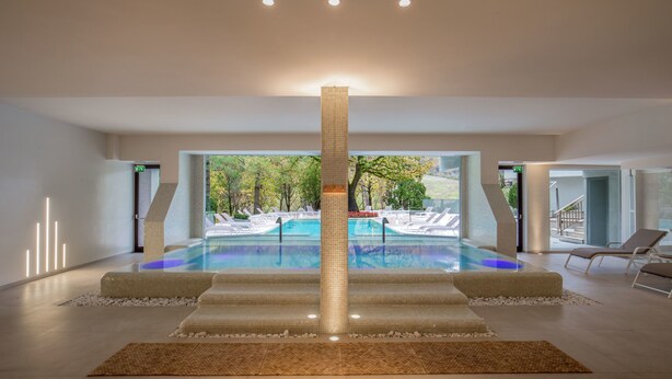 Un área de piscina interior moderna con un pilar central, sillas de descanso y vistas al exterior.