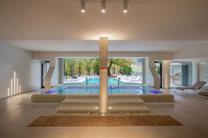 Un área de piscina interior moderna con un pilar central, sillas de descanso y vistas al exterior.