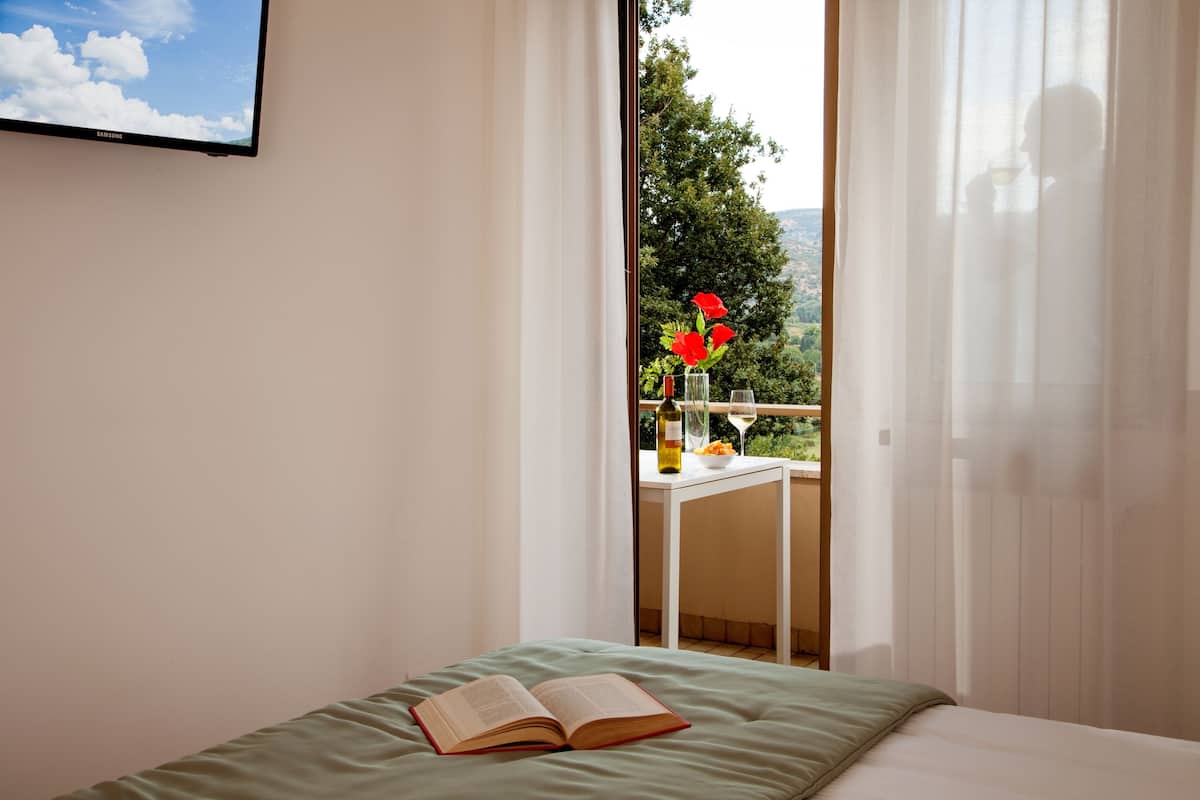 Un dormitori amb un llit, un llibre obert, una finestra amb cortines translúcides, i una vista a vegetació i una taula amb una ampolla i flors a l'exterior.