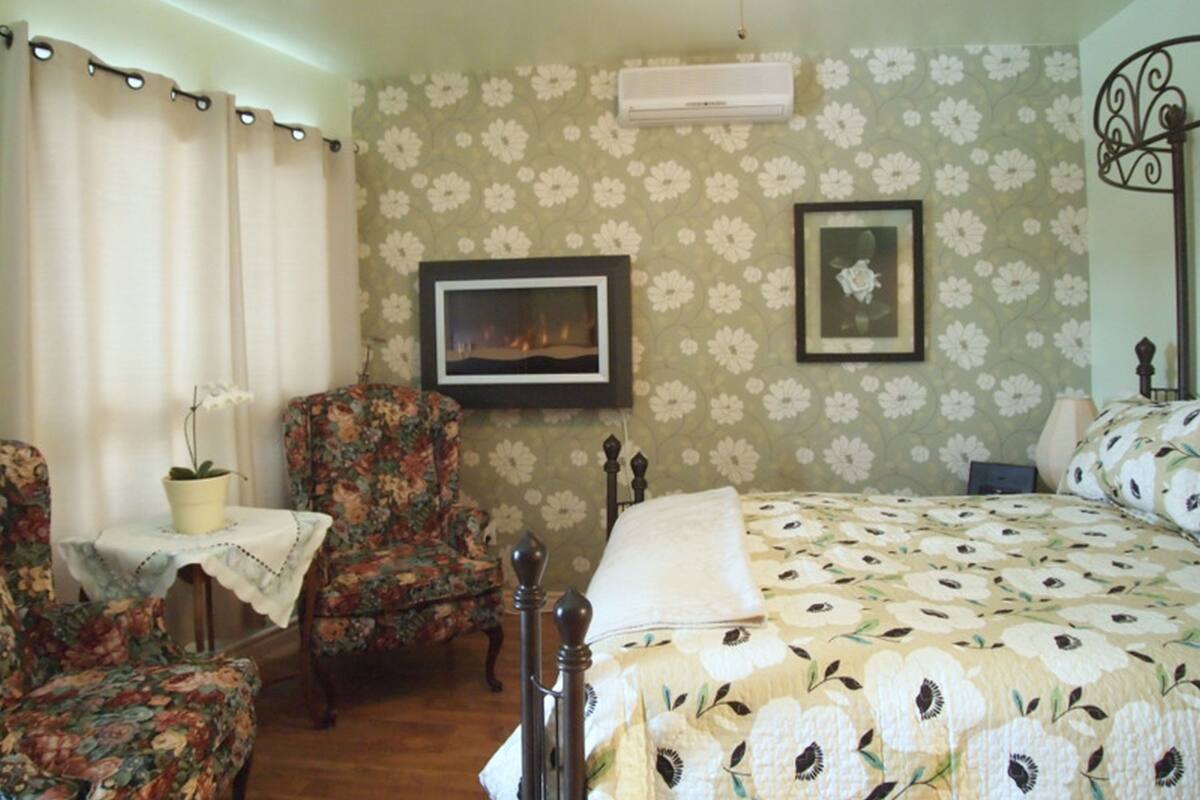 Un dormitorio con papel tapiz floral, una cama con colcha estampada, una silla, una mesa con planta en maceta y un ventilador de techo.