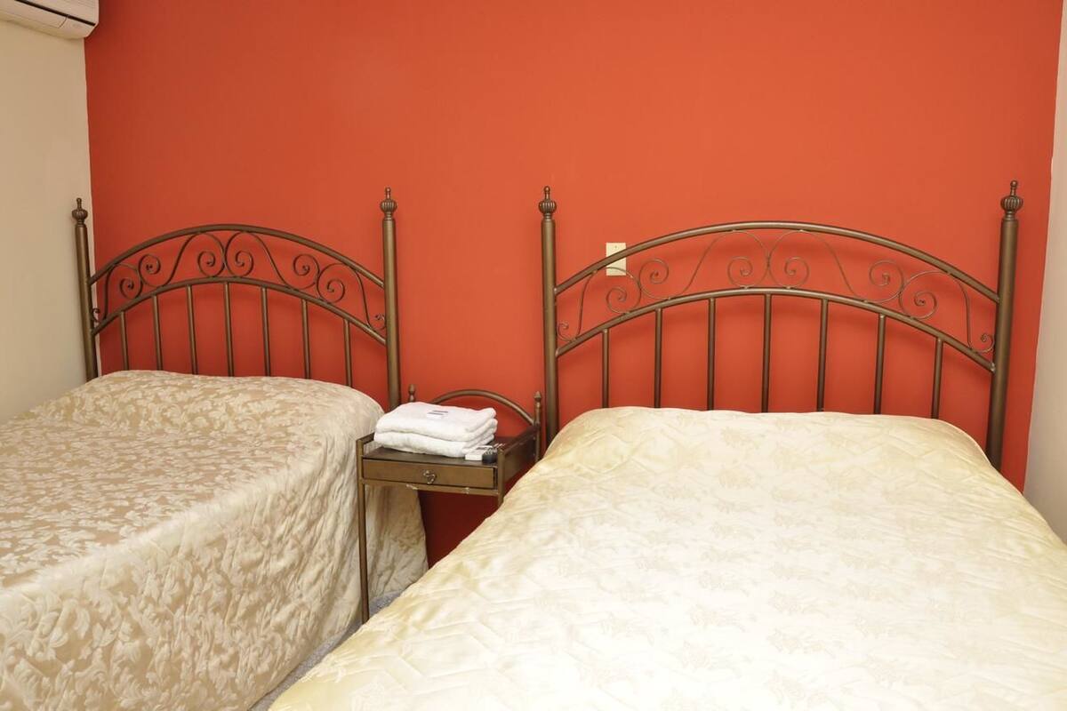 A room with two beds, each with a metal headboard and a beige bedspread. There is a small wooden nightstand between the beds with a folded white towel on it.
