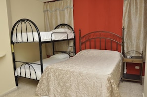A bunk bed with a patterned bedspread, a bedside table with a lamp, and a red wall. - Hotel Monumental (Santo Domingo)