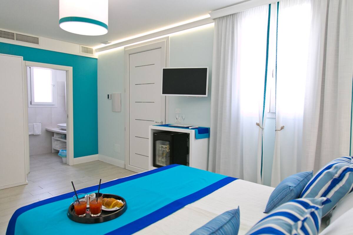 A hotel room with a bed, a TV mounted on the wall, a bathroom visible through an open door, and a tray with drinks on the bed.