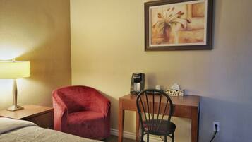 A hotel room with a bed, a red chair, a desk with a coffee maker, a framed painting, and a lamp.