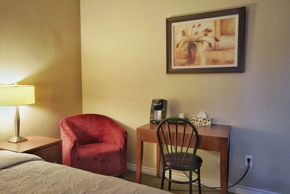 Habitación de hotel con una cama, una silla roja, un escritorio con cafetera, un cuadro enmarcado y una lámpara.