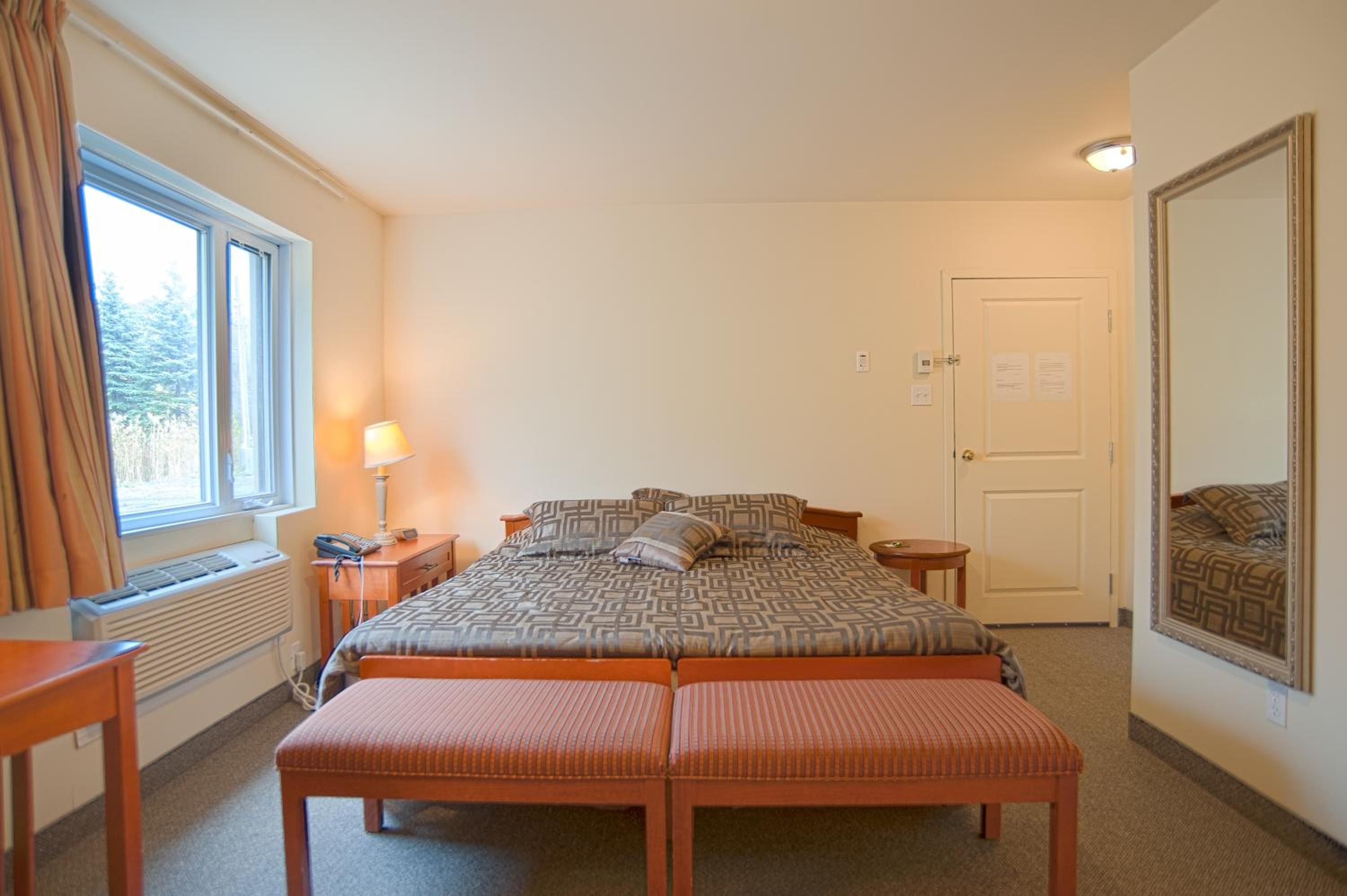 Una habitación de hotel con cama, mesita de noche, lámpara, ventana, puerta y espejo.
