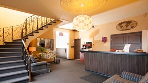 Un vestíbulo de hotel con una escalera, un mostrador de recepción, un sofá estampado y una lámpara decorativa en el techo.