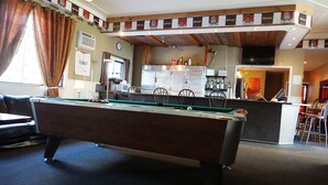 Una mesa de billar en una sala con área de bar, un reloj en la pared y una ventana con cortinas.