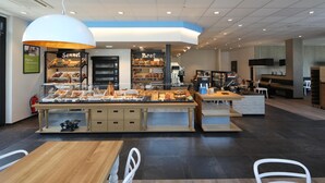 Mostrador de panadería con diversos productos horneados a la vista, zona de estar con mesas y sillas, y mostrador con máquina de café.