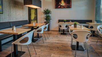 A restaurant with wooden tables and white chairs, a fire feature on the wall, and a potted plant.