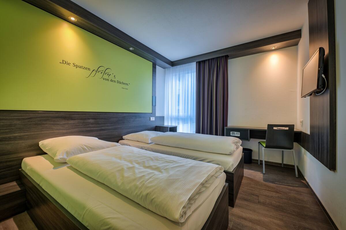 Habitación de hotel con dos camas, un escritorio, una silla, un televisor y una pared verde grande con texto.