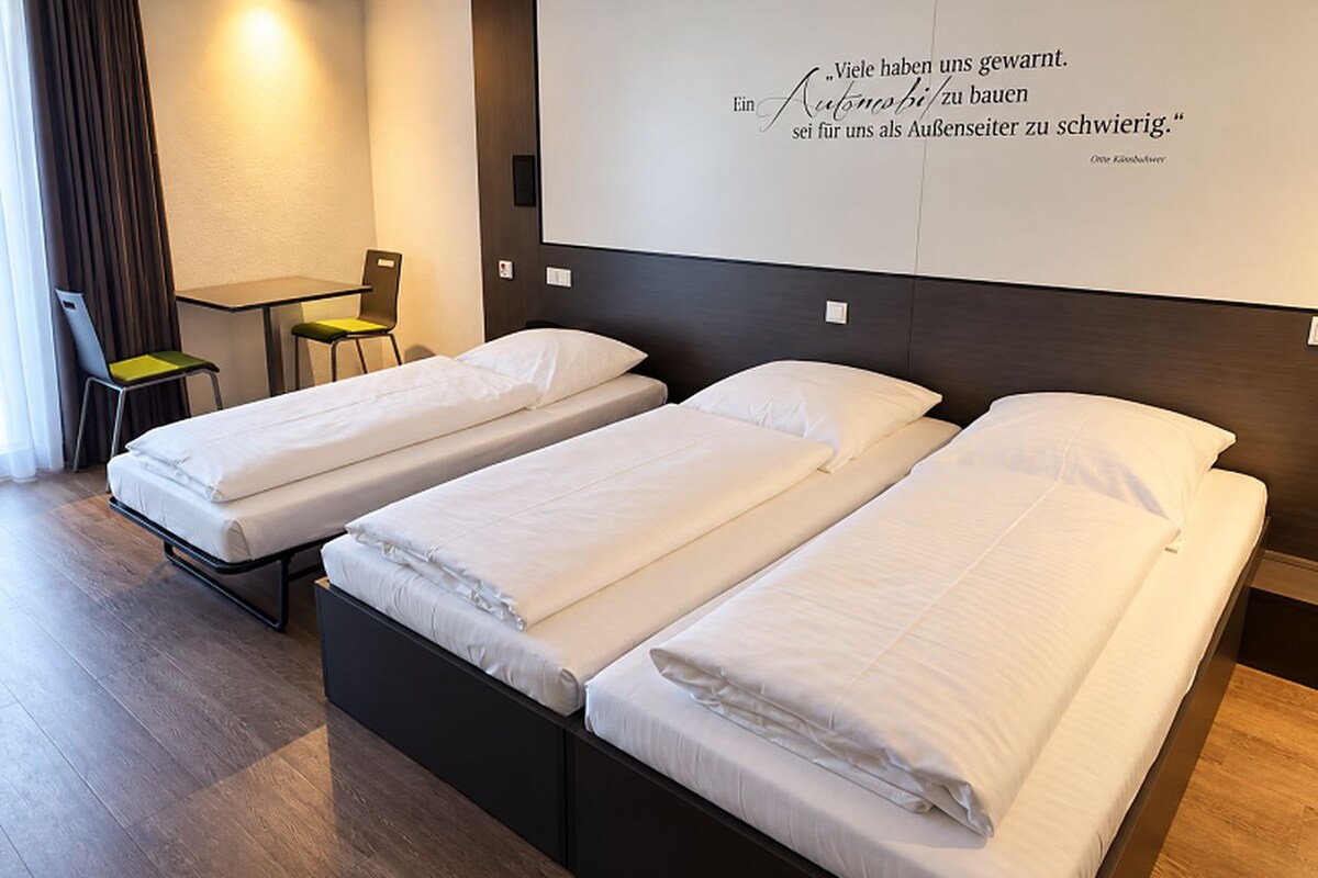 Habitación de hotel con tres camas individuales, una mesita con dos sillas y una pared con una cita.
