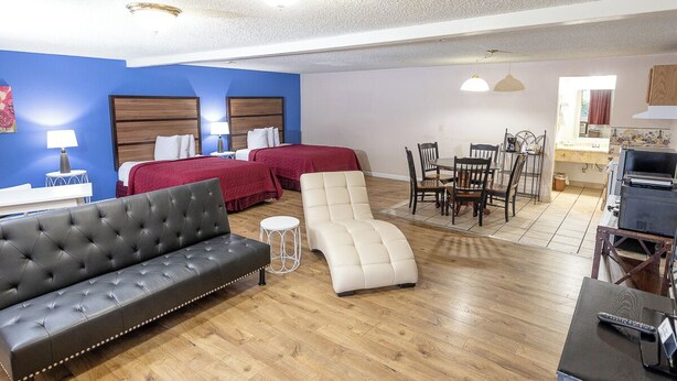 Habitación de hotel con sofá de cuero negro, sillón reclinable blanco, dos camas con ropa de cama roja, una mesa de comedor con sillas y una zona de kitchenette.