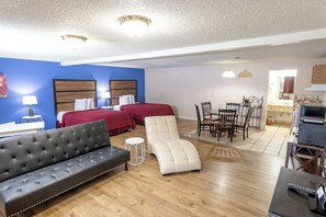 Habitación de hotel con sofá de cuero negro, sillón reclinable blanco, dos camas con ropa de cama roja, una mesa de comedor con sillas y una zona de kitchenette.