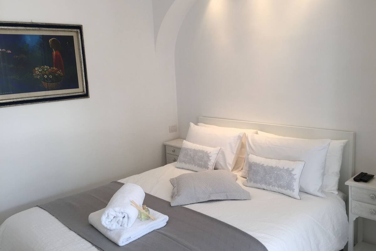 A neatly made bed with white bedding and a grey throw, a nightstand with a phone, and a framed picture on the wall.